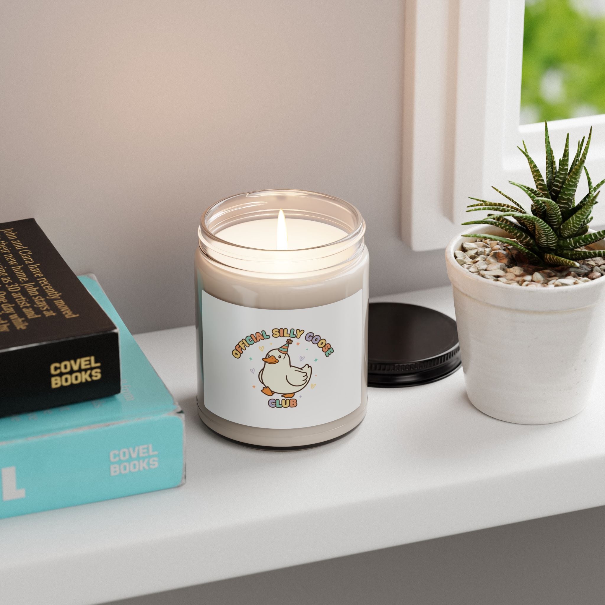 Candle with a visible brand logo on a shelf with books and a plant